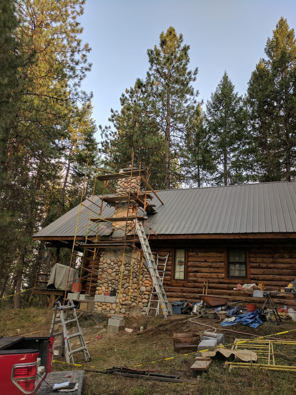 manufactured river rock chimney built past the metal roof to the crown with scaffold still standing against the cabin