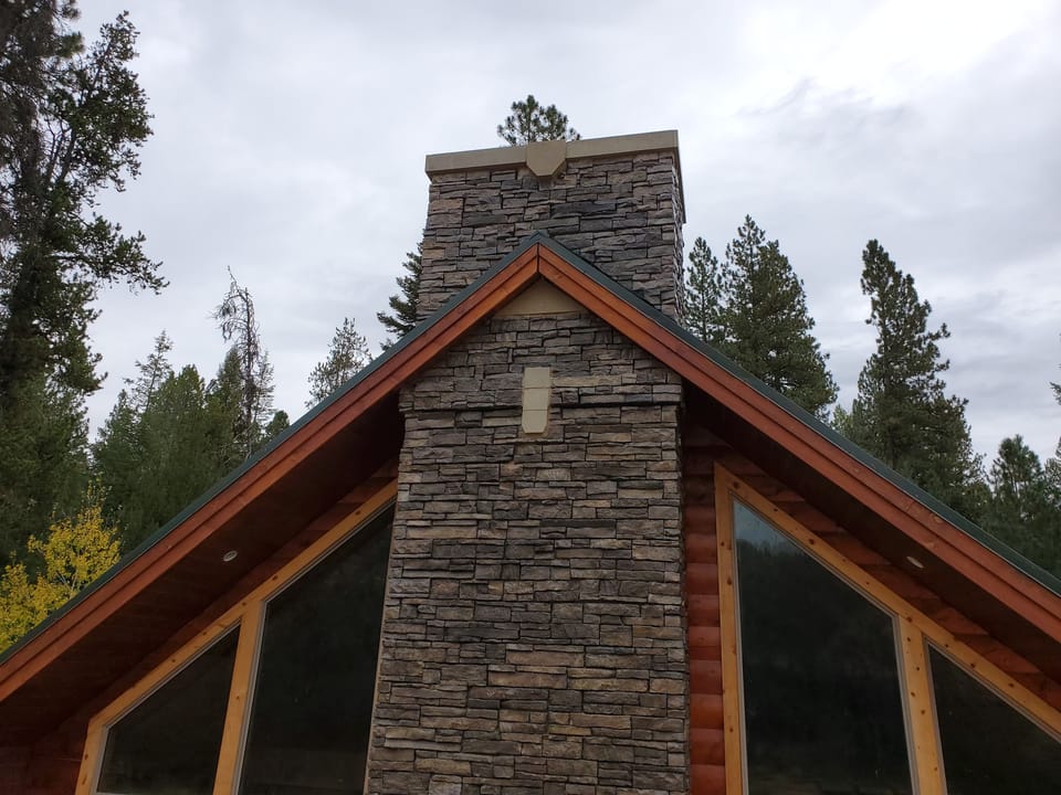 Finished stone veneer chimney with a cast stone crown, seen from below against pines and sky